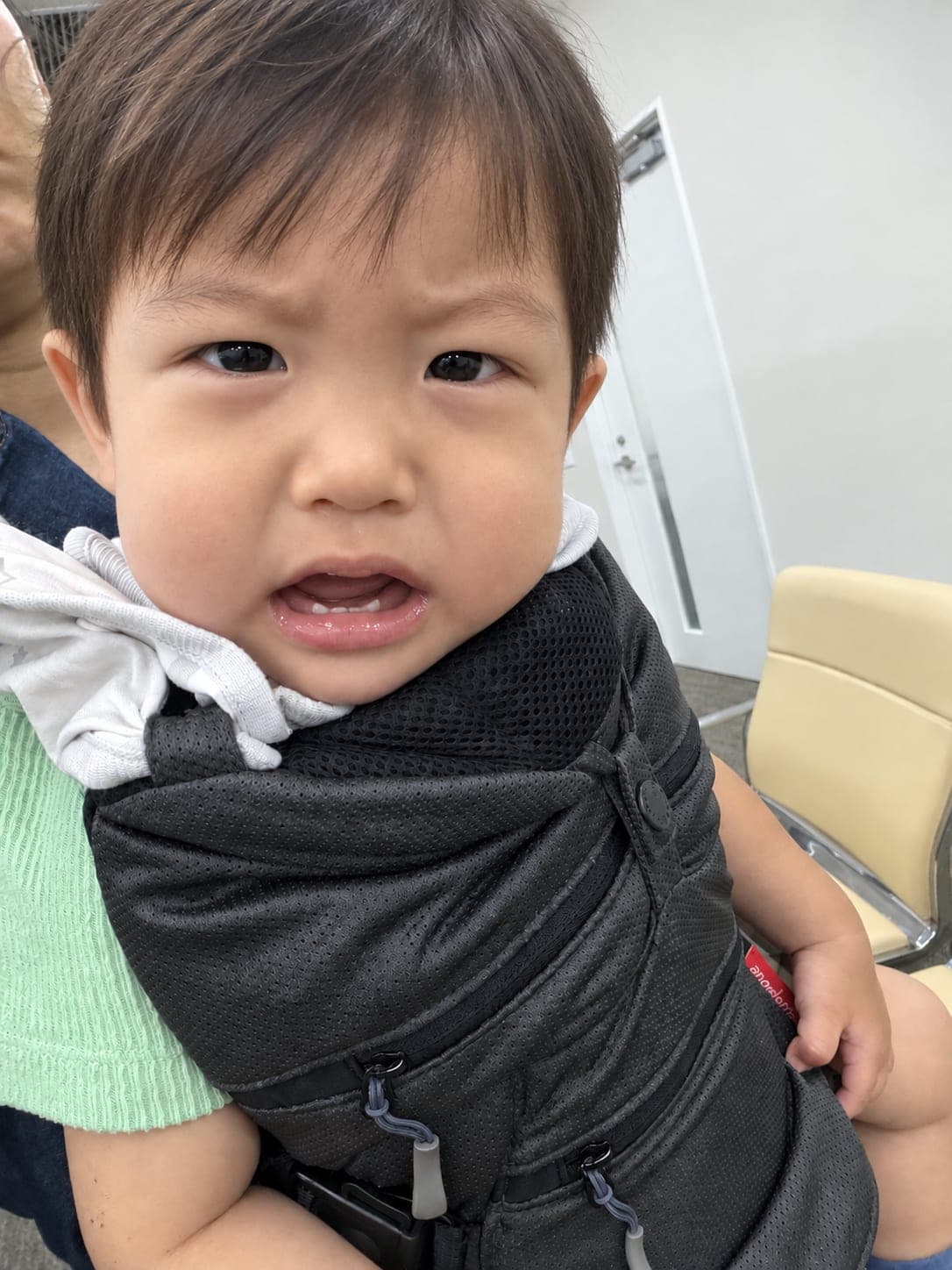 抱っこ紐の中で眉をひそめて泣きそうな顔をしている1歳の子ども