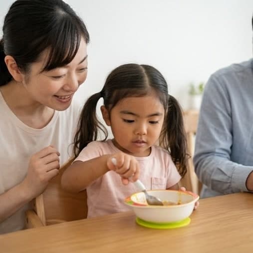 お母さんに見守られて、自分でスプーンを使って食べようとしている女の子