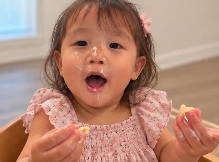 口のまわりを食べこぼしで白くしながら、自分の手でおやつを食べている女の子