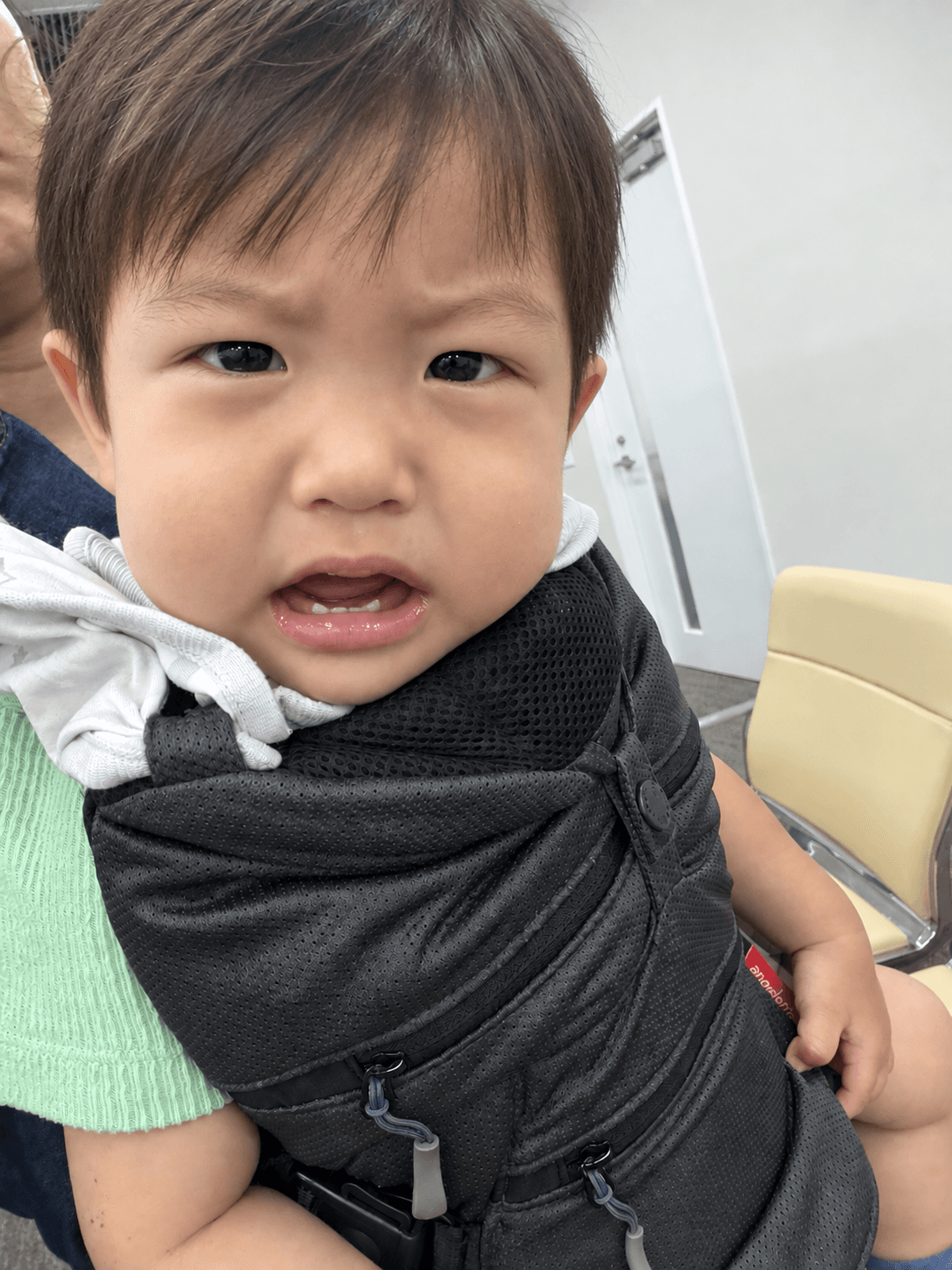 抱っこ紐の中で眉をひそめて泣きそうな顔をしている1歳の子ども